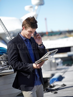 Buy stock photo Young businessman reading the newspaper while on his cellphone at the marina
