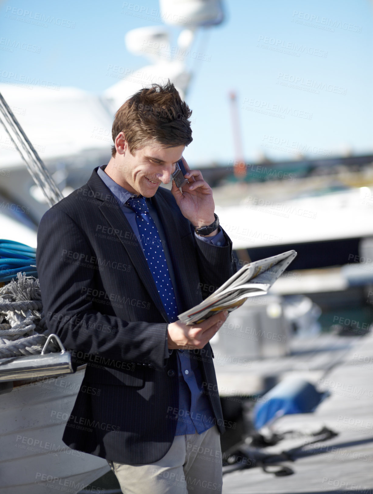 Buy stock photo Young businessman reading the newspaper while on his cellphone at the marina