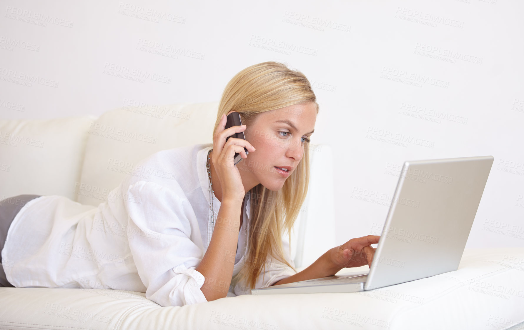 Buy stock photo A gorgeous young woman working on her laptop while lying on the sofa and talking on her cell