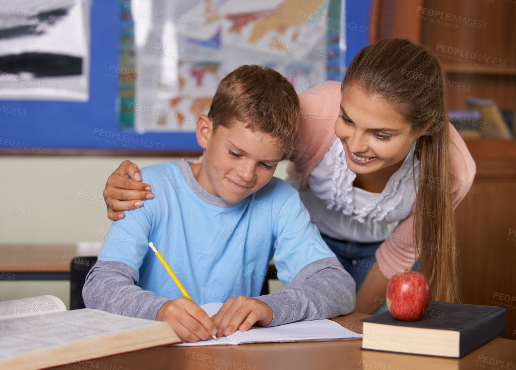 Buy stock photo A supportive young teacher leaning over her student while he writes in a book
