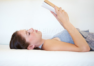 Buy stock photo A young woman reading a book while lying on the couch