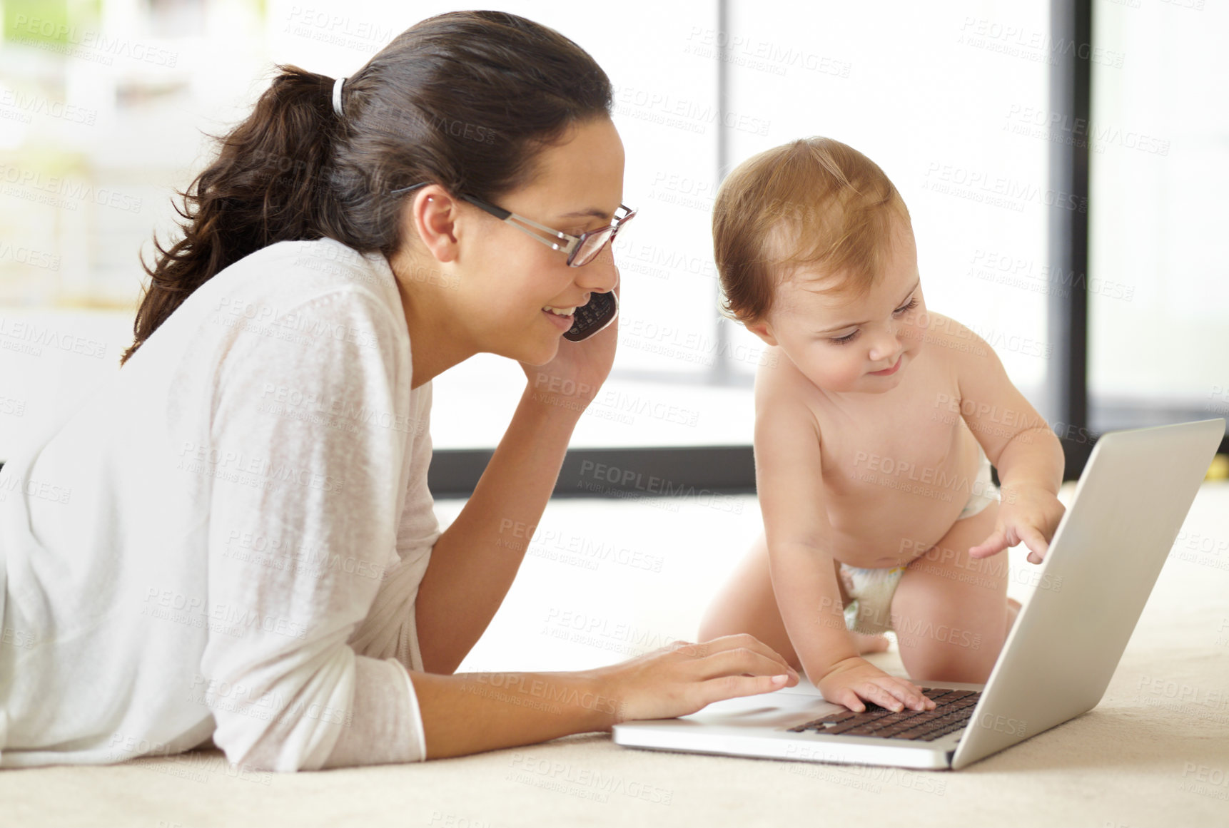 Buy stock photo A young mother speaking on the phone while lying with her baby in front of an open laptop