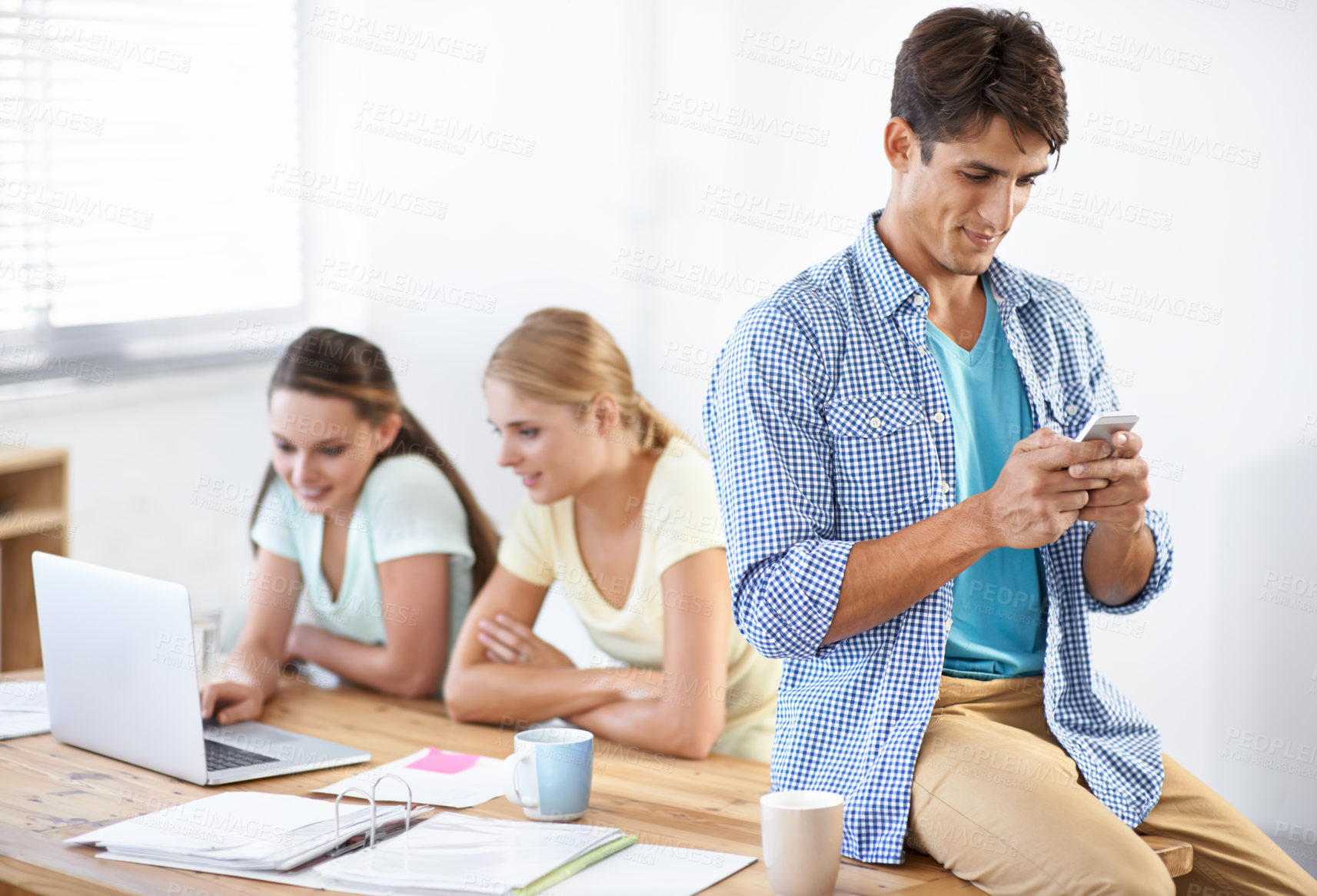 Buy stock photo A handsome young man typing on his phone while his colleagues work on a laptop together