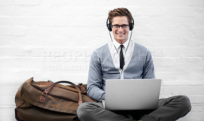 Buy stock photo A handsome young business man sitting on the ground wearing headphones while typing on his laptop with luggage next to him
