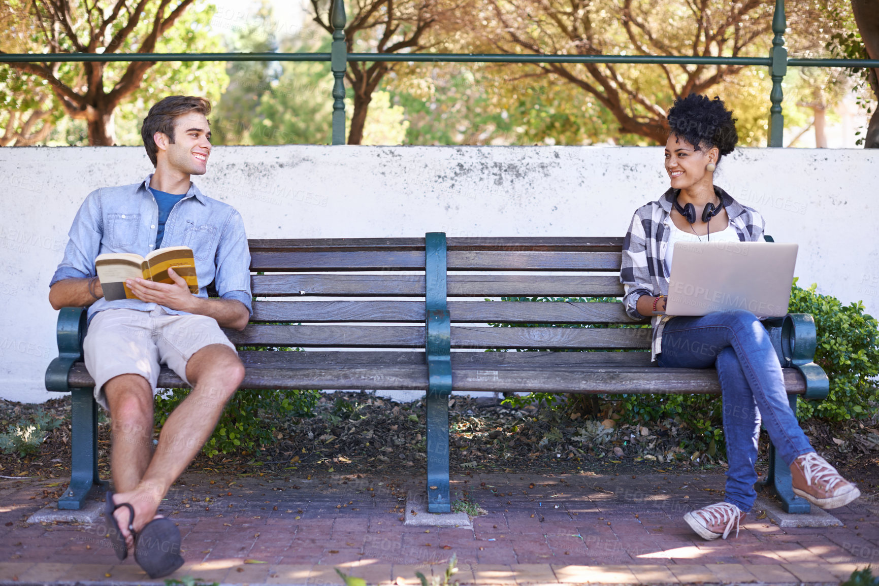 Buy stock photo Two students sitting on opposite sides of a bench with their reading material in front of them and looking at each other