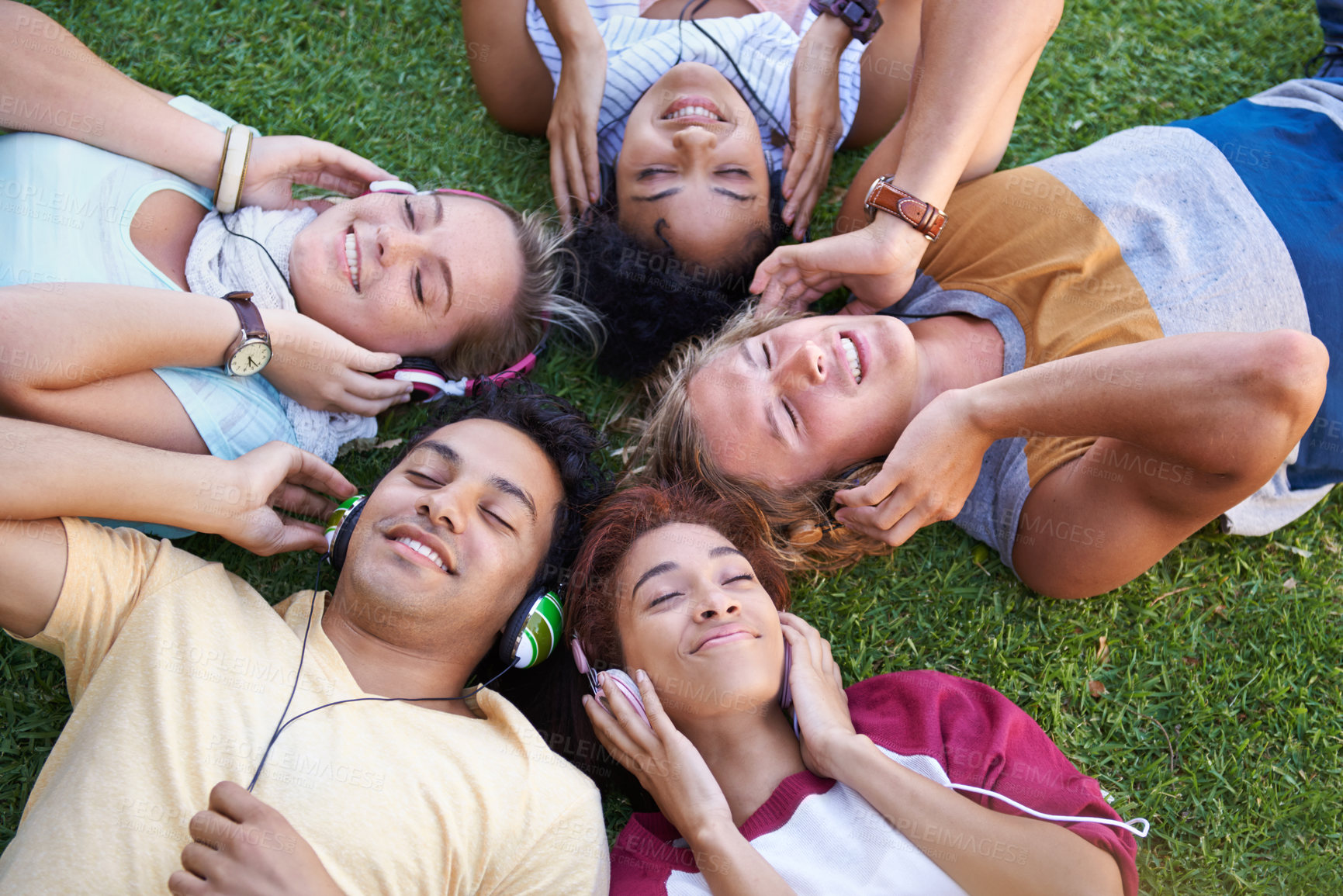 Buy stock photo High angle of a group of friends lying on their backs on the grass in a park and listening to music