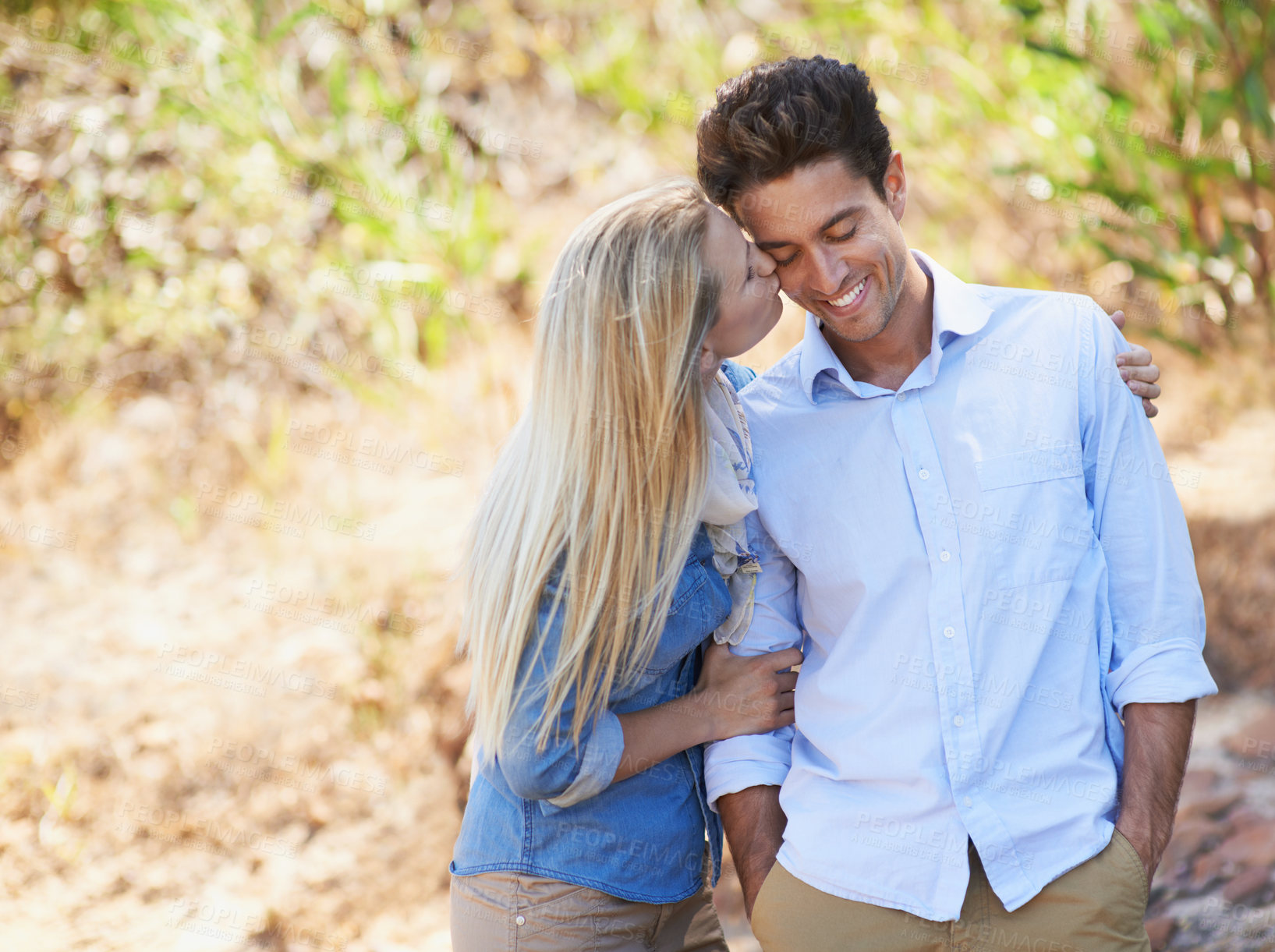 Buy stock photo Happy couple, kiss and hug in nature for love, support or affection in outdoor walk or bonding. Young woman and man on cheek with smile for embrace, comfort or romance in forest or woods together