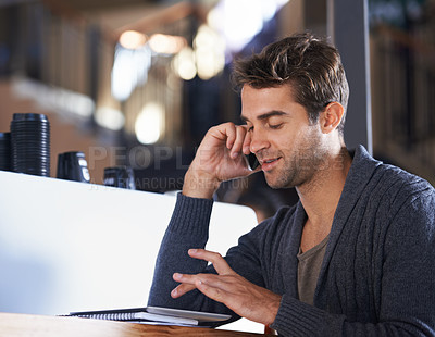 Buy stock photo A young man sitting in a coffee shop speaking on his cellphone and working on a digital tablet