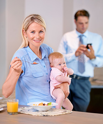 Buy stock photo Mid-aged mother having a healthy breakfast , while holding her baby and sitting at the kitchen table. While father is on a business texting , in the background and working hard for his family 