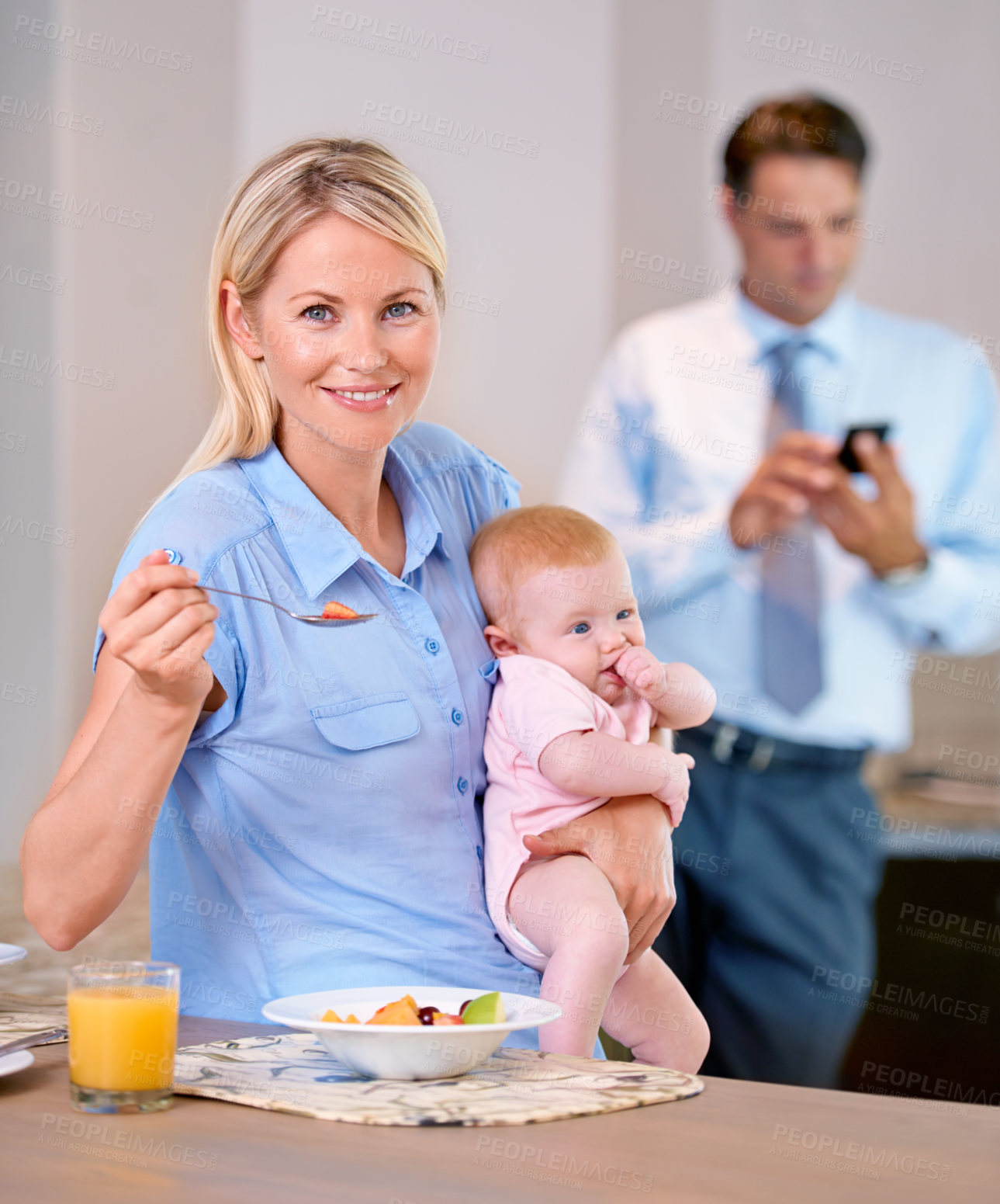 Buy stock photo Mid-aged mother having a healthy breakfast , while holding her baby and sitting at the kitchen table. While father is on a business texting , in the background and working hard for his family 