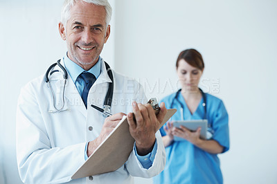Buy stock photo Portrait, mature doctor writing on his clipboard and with a nurse browsing on a digital tablet in the background. Senior doctor and nurse standing in corridor looking at files discussing reports