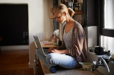 Buy stock photo An attractive young woman using her laptop while sitting on her kitchen counter