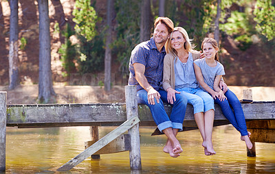 Buy stock photo A happy family of three sitting together on the jetty at the lake