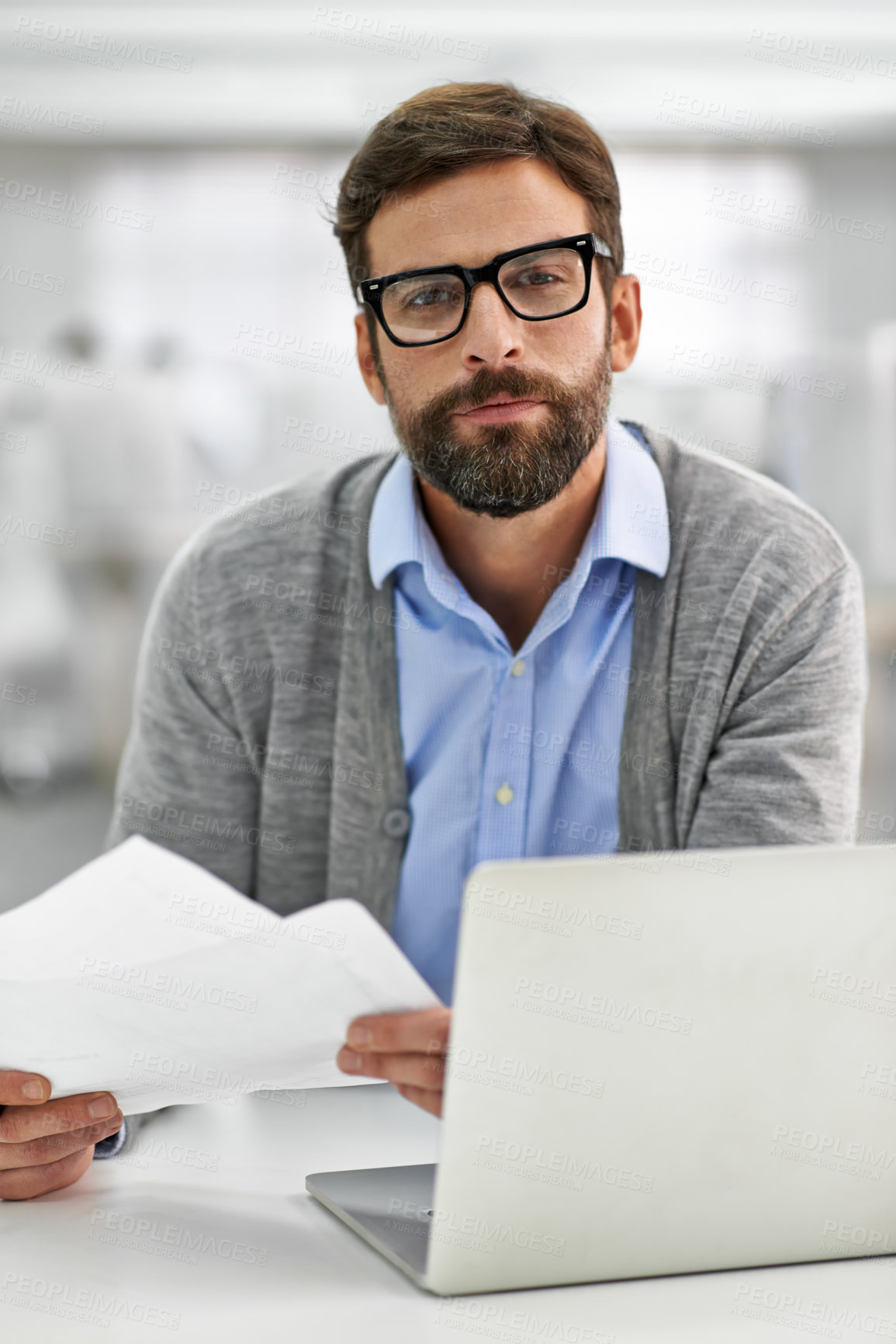 Buy stock photo A young businessman going over paperowrk while working on his laptop in the office