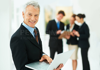 Buy stock photo Portrait of a mature businessman holding a laptop with colleagues in the background