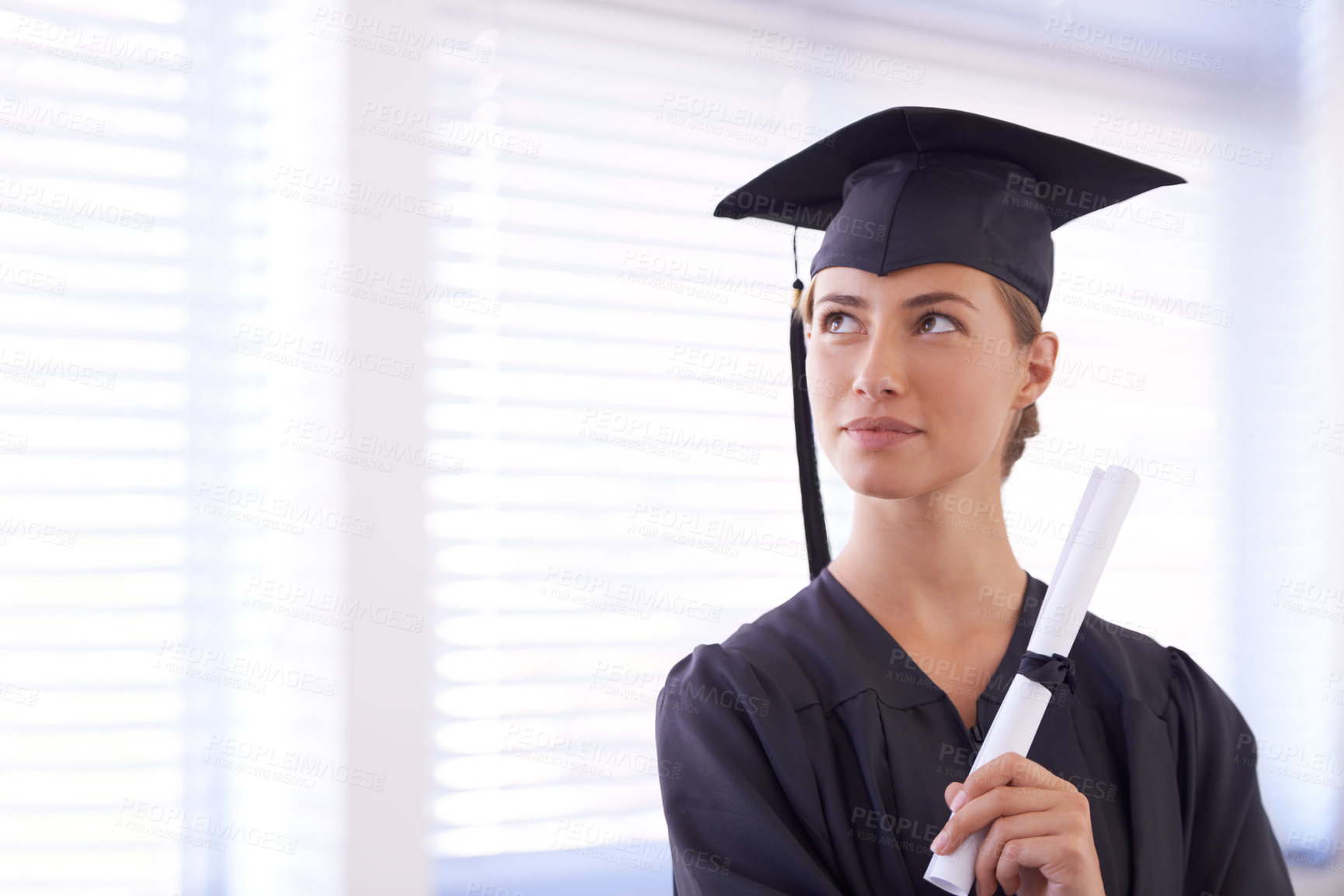 Buy stock photo Shot of a young woman in a graduation gown holding a diploma and looking off to the side