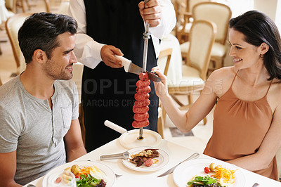 Buy stock photo Couple, restaurant and waiter serving food or sausage kebab on skewer for fine dining, anniversary or nutrition. Man, woman and relationship bonding dates in Italy with water carving, meal or eating