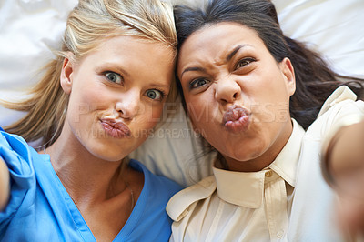 Buy stock photo High angle portrait of two attractive young women taking selfies while lying on a bed