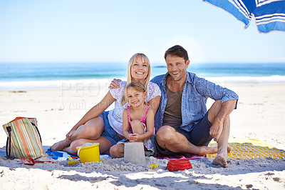 Buy stock photo Parents, girl and sand castle in portrait by ocean, blanket and umbrella with hug on holiday in summer. Father, mother and daughter with picnic at beach for vacation in nature with love in sunshine