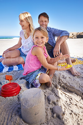 Buy stock photo Parents, child and sandcastle in portrait at beach, blanket and excited with smile for holiday in summer. Father, mother and daughter with family picnic by ocean for vacation with love in sunshine