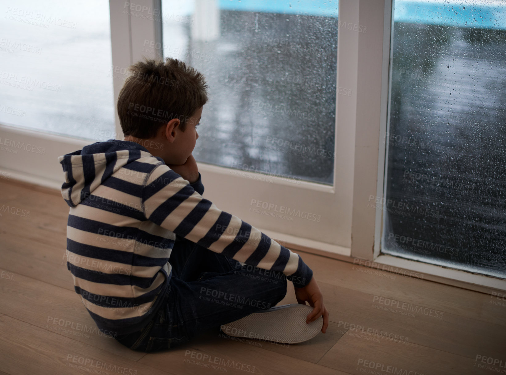 Buy stock photo Rain, window and sad boy child on a floor unhappy, upset or annoyed by bad weather in his home. Glass, depression and kid in a living room frustrated by storm, cold or unexpected winter disaster