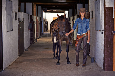 Buy stock photo Shot of a cowgirl leading a horse out of a stable
