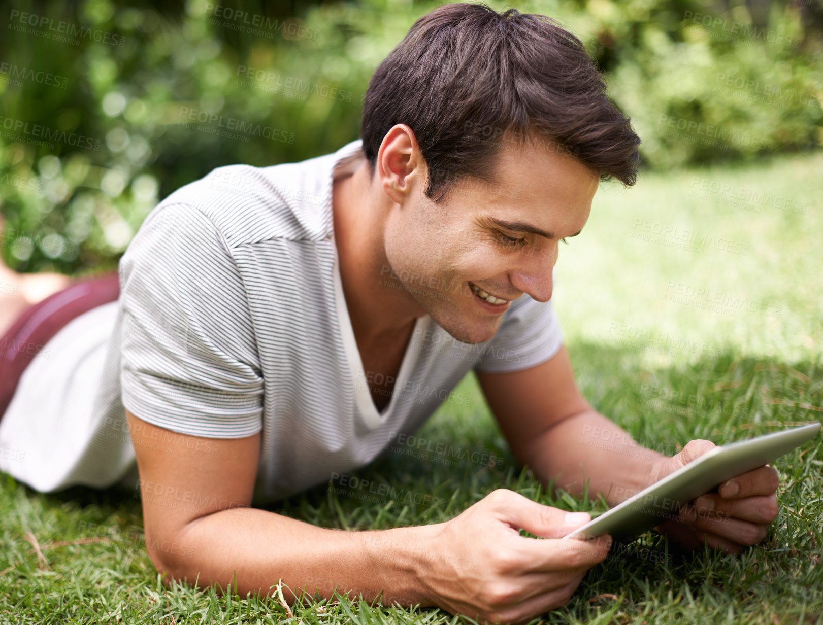 Buy stock photo Shot of a handsome young man using a digital tablet while lying outdoors