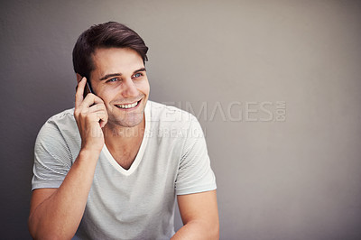 Buy stock photo Handsome young man talking on his phone and smiling while sitting against an isolated grey background with copyspace. Happy guy calling his friends to share great news about his phone subscription 