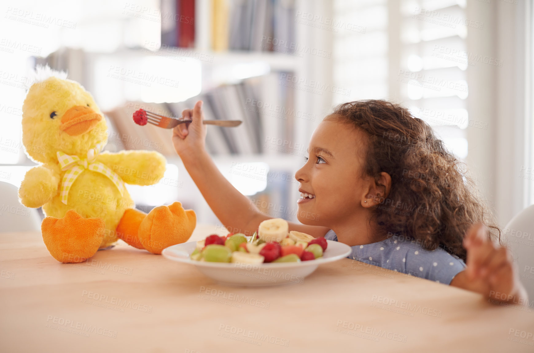 Buy stock photo Child feeding, teddy bear and food at home with happiness and wellness meal with a smile. House, nutrition and young girl with healthy fruit salad and fruits of a hungry kid with toy duck at a table 
