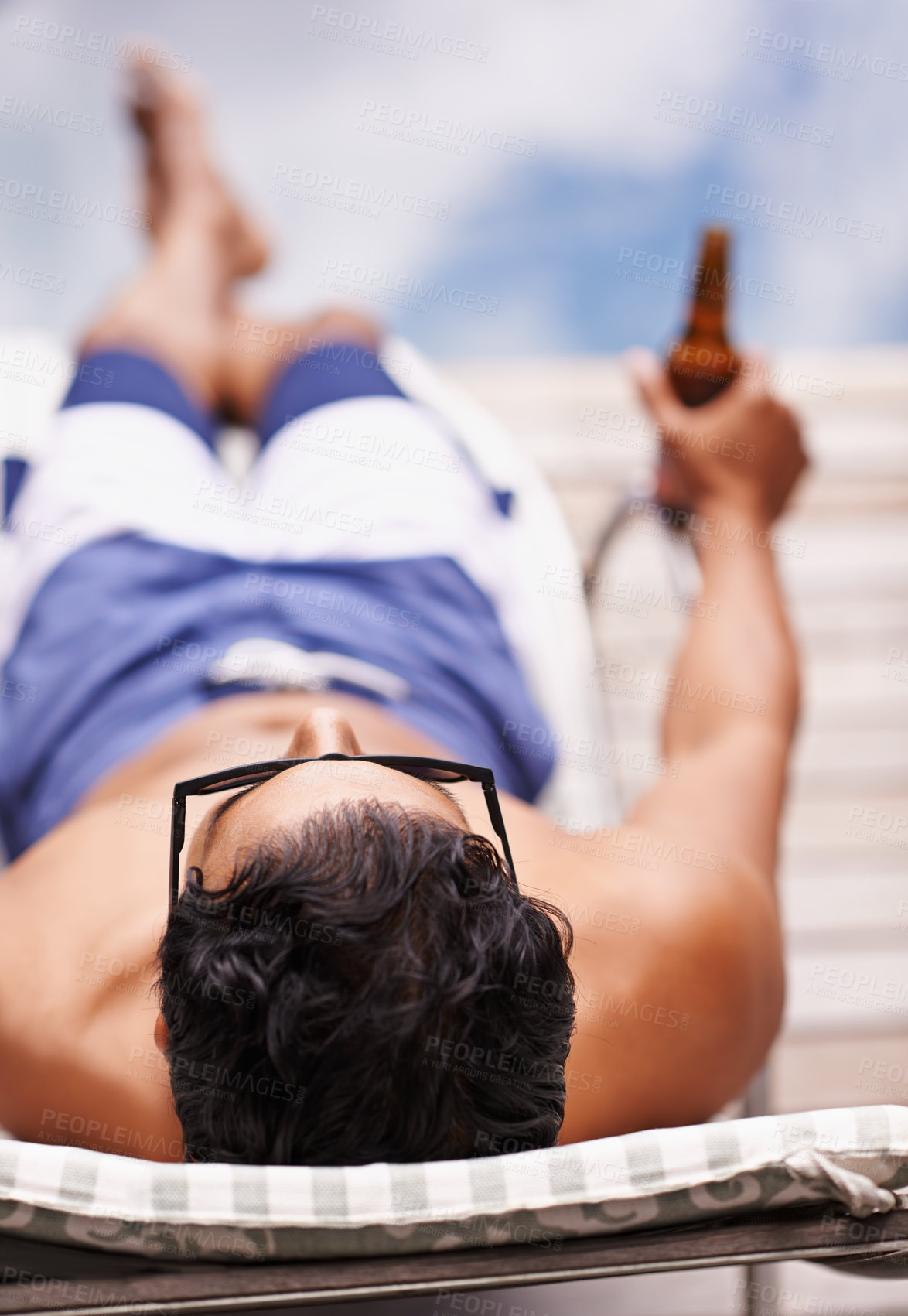 Buy stock photo Rear view shot of a young man lounging be the pool with a beer