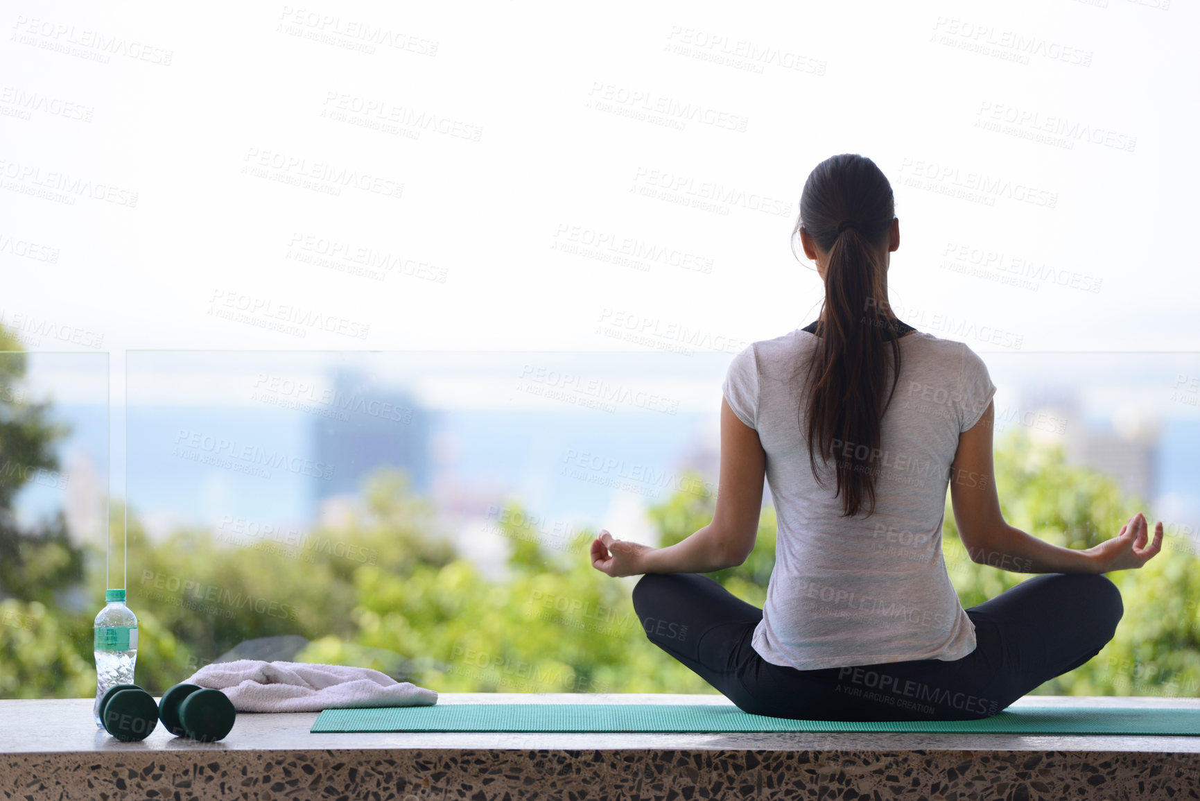 Buy stock photo A young woman meditating on a yoga mat by a window overlooking the city