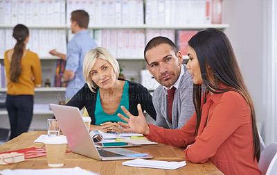 Buy stock photo Three coworkers brainstorming around a laptop while their colleagues read files in the background