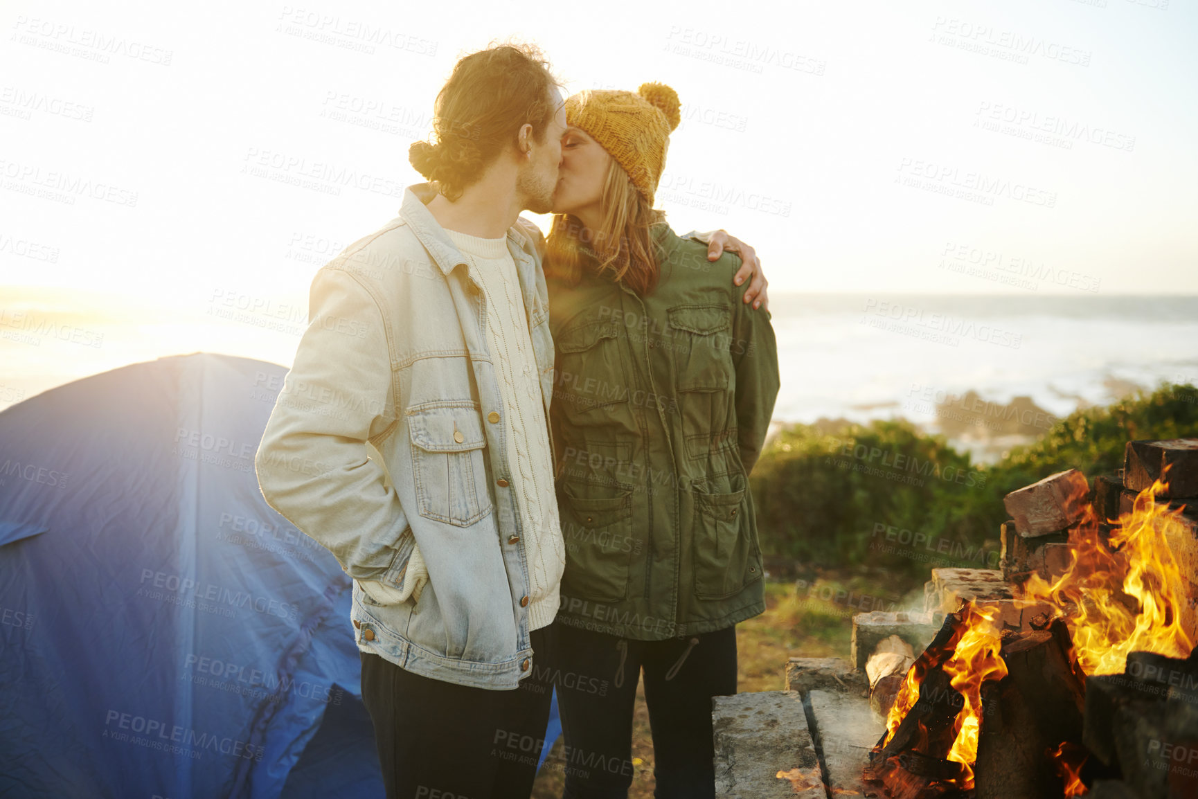 Buy stock photo Camping, fire and kissing with couple on beach together for holiday, romance or vacation in summer. Nature, sunset or tent with young man and woman hugging at coast by ocean or sea for adventure