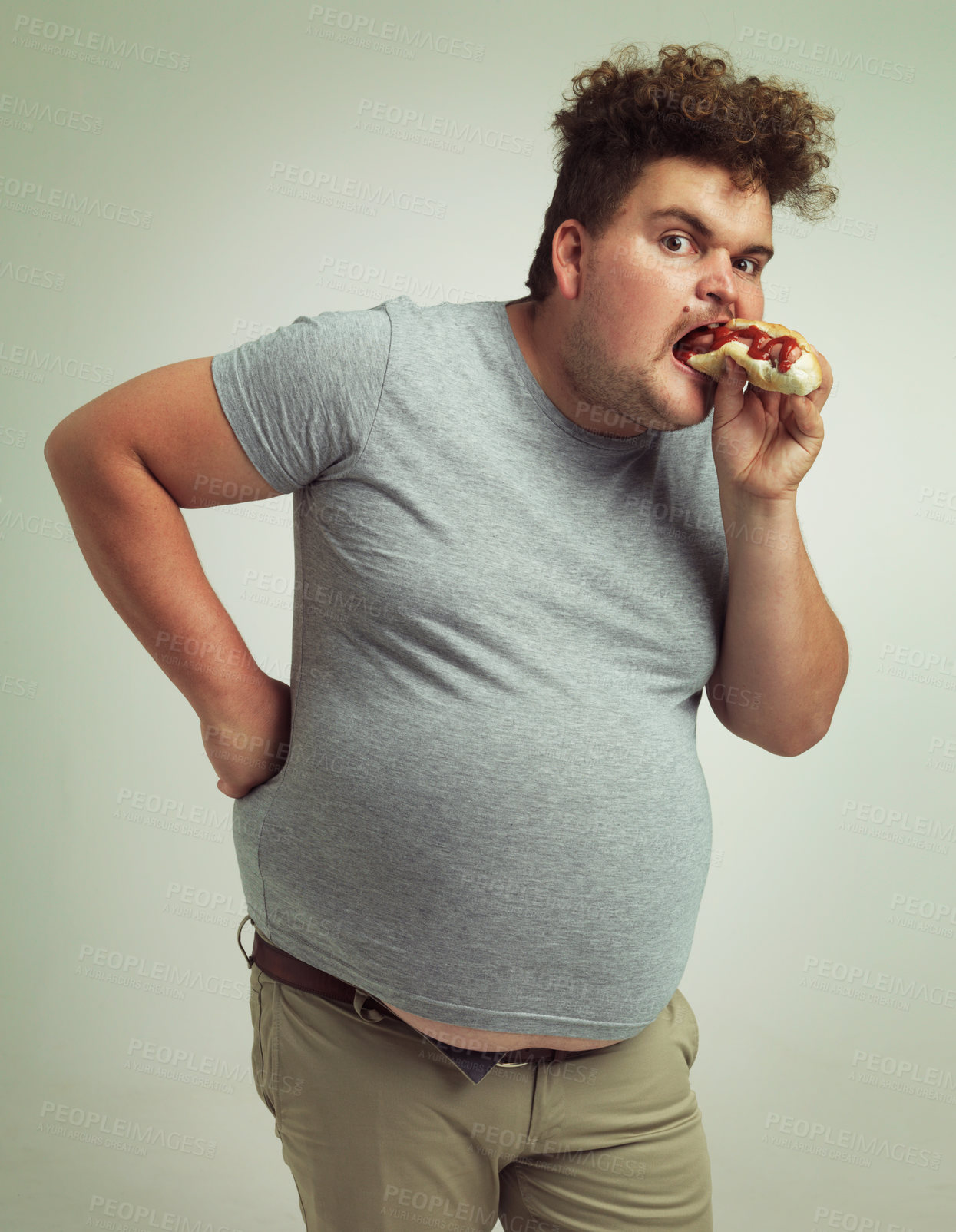 Buy stock photo Man, fast food and eating hotdog for lunch in studio background for snack, hunger and craving for plus size guy. Male person, hungry and burger or takeaway meal for starving and enjoy while standing