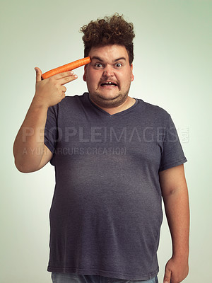 Buy stock photo Portrait, vegetable and hand gun with plus size man in studio on gray background for diet, health or nutrition. Anger, depression and carrot to head of young person in conflict with vegetarian food