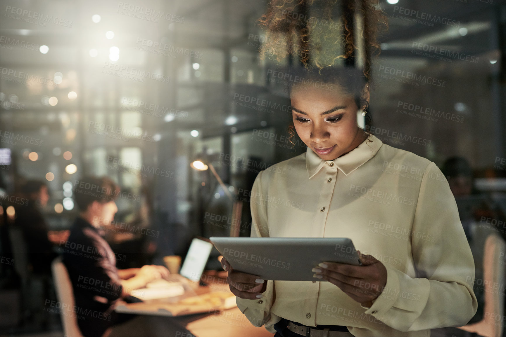 Buy stock photo Shot of an attractive young designer working late at the office with her colleagues in the background