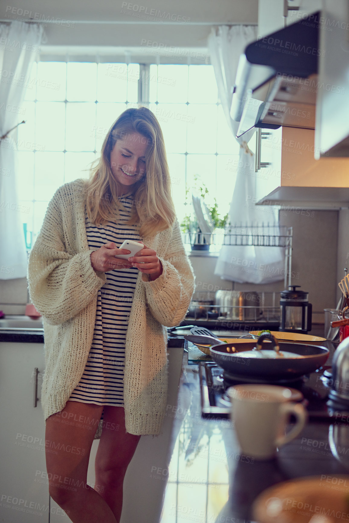 Buy stock photo Cropped shot of an attractive young woman texting on a cellphone while cooking in her kitchen