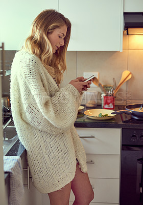 Buy stock photo Cropped shot of an attractive young woman texting on a cellphone while cooking in her kitchen
