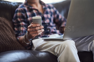 Buy stock photo Shot of an unrecognizable man using his laptop and cellphone while sitting on the sofa at home