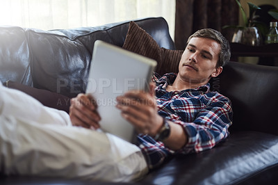 Buy stock photo High angle shot of a handsome young man using his tablet while sitting on the sofa at home