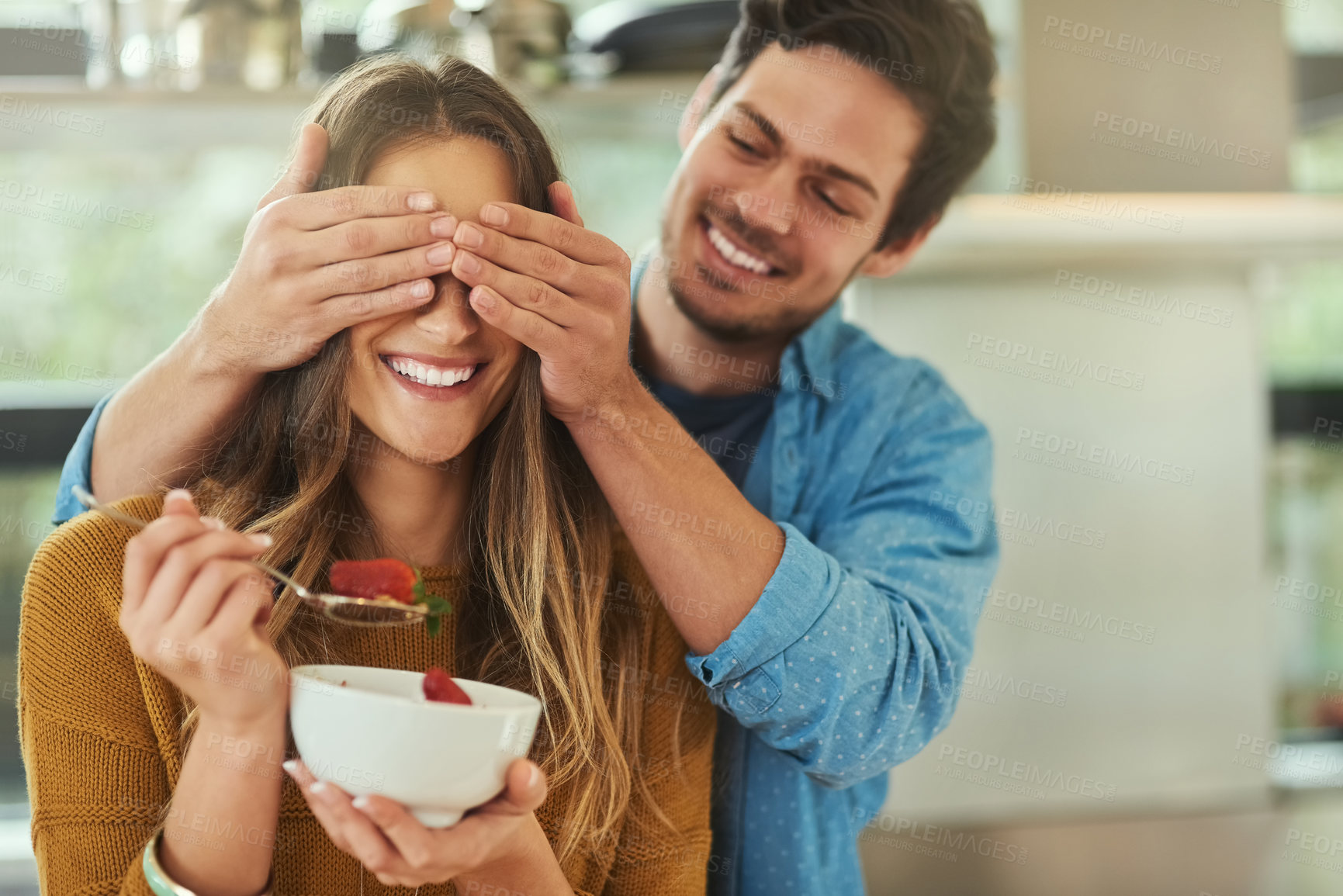Buy stock photo Shot of an affectionate young man covering his girlfriend's eyes while she eats breakfast in their kitchen