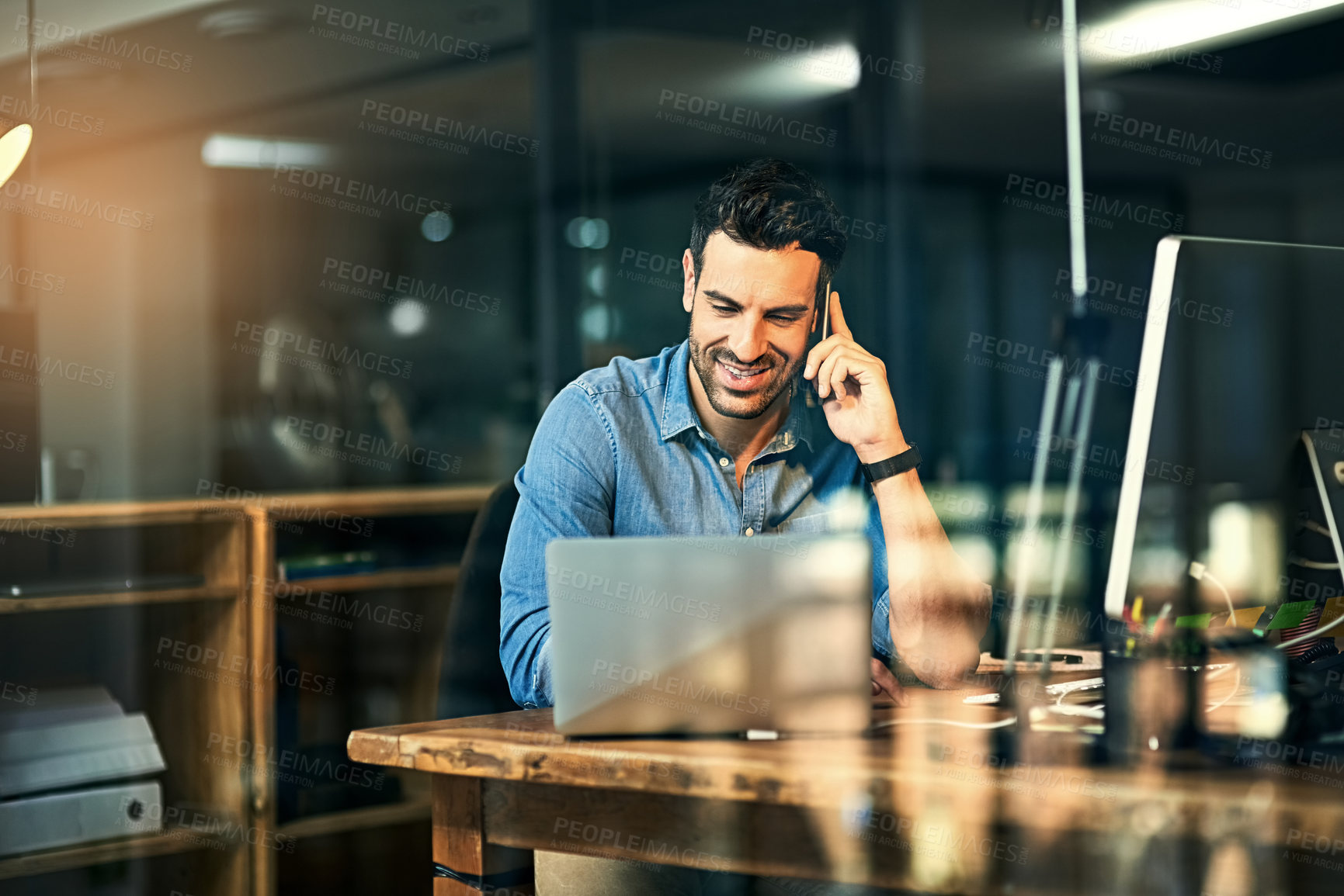 Buy stock photo Shot of a young businessman talking on his phone and using a laptop during a late night at work