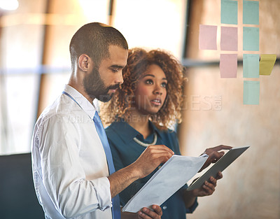 Buy stock photo Shot of  two young professionals brainstorming with sticky notes on a glass wall in an office