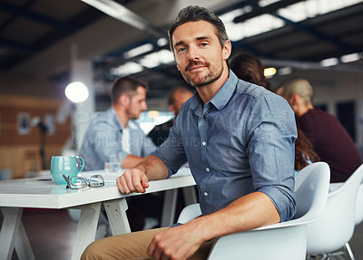 Buy stock photo Portrait of a mature man sitting at a table in an office with colleagues working in the background