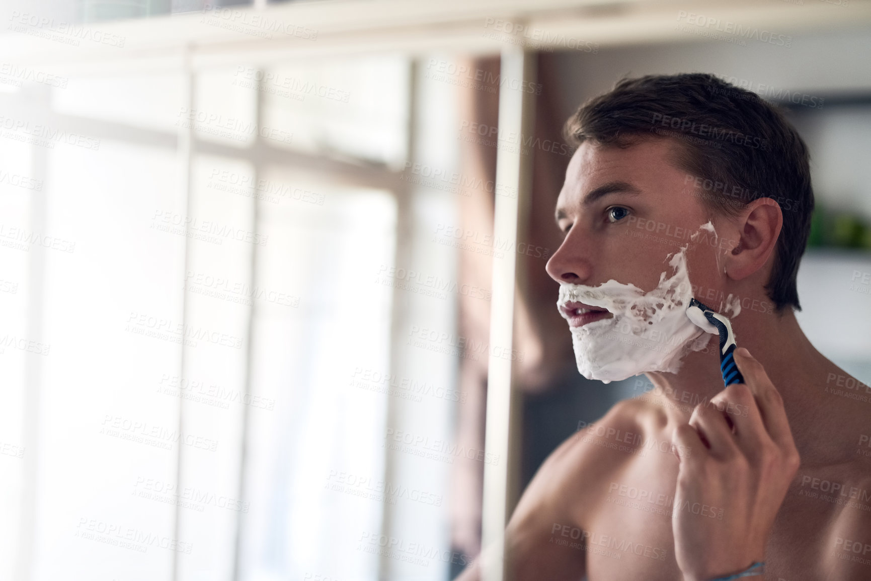 Buy stock photo Shot of a young man shaving his beard while looking at himself in the mirror