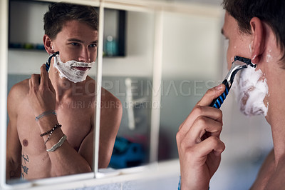 Buy stock photo Shot of a young man shaving his beard while looking at himself in the mirror