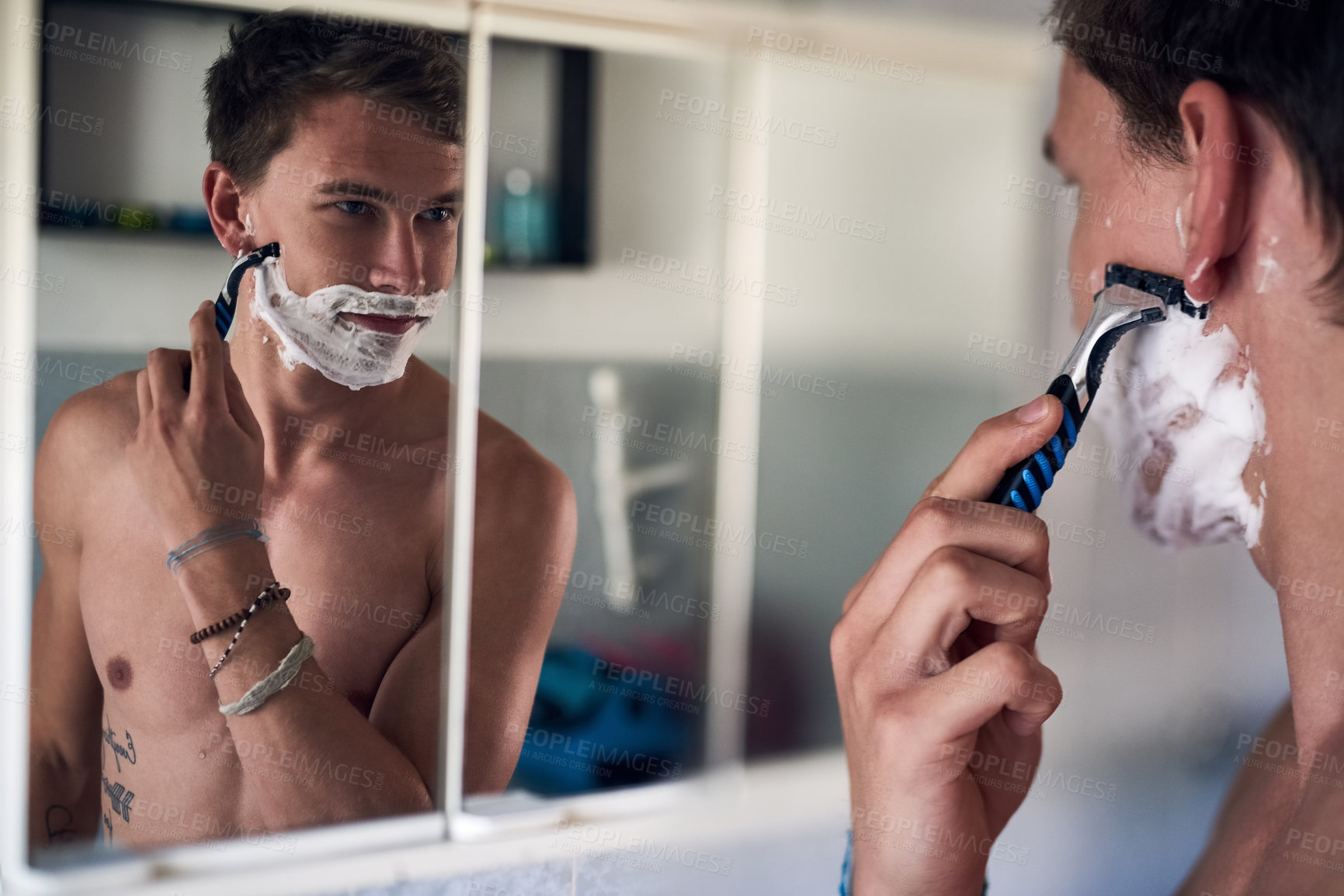 Buy stock photo Shot of a young man shaving his beard while looking at himself in the mirror