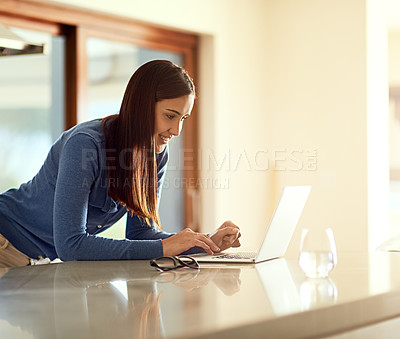 Buy stock photo Shot of a happy young woman using her laptop while standing in her kitchen