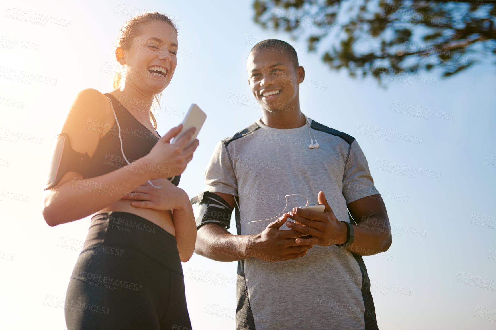 Buy stock photo Fitness, couple and phone with music outdoor for exercise, radio app and laughing on workout break in park. Interracial people, earphones and smartphone in nature for audio playlist, bonding and rest