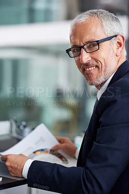 Buy stock photo Portrait of a smiling mature businessman reading paperwork while sitting at his desk in an office